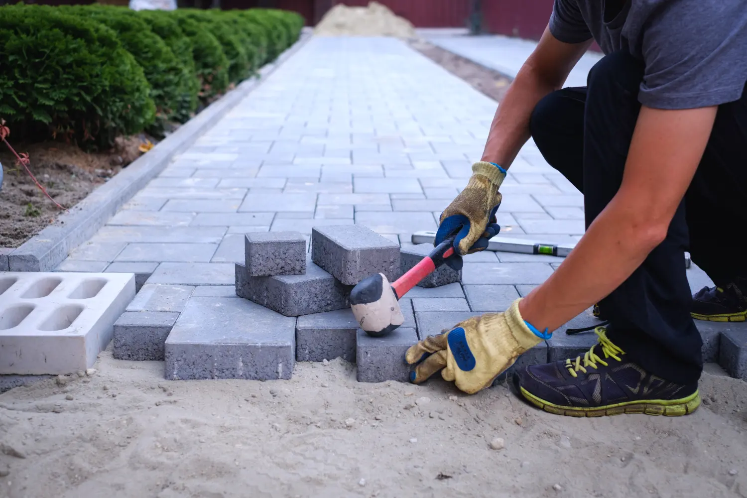 Der Meister mit den gelben Handschuhen verlegt schichtweise Pflastersteine. Pflasterung eines Gartenwegs mit Ziegelsteinen durch einen professionellen Pflasterer. 